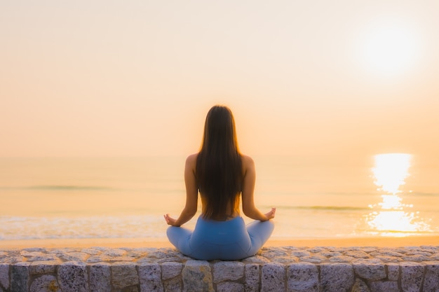 La giovane donna asiatica del ritratto fa la meditazione intorno all'oceano della spiaggia del mare all'alba