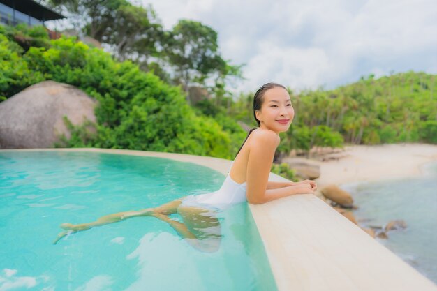 La giovane donna asiatica del ritratto si rilassa il sorriso felice intorno alla piscina all'aperto nella località di soggiorno dell'hotel con la vista di oceano del mare