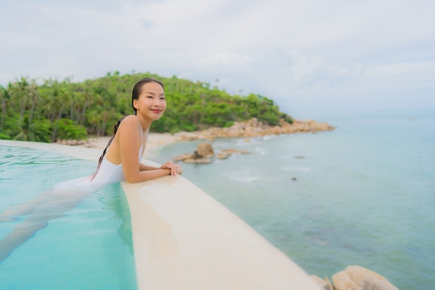 La giovane donna asiatica del ritratto si rilassa il sorriso felice intorno alla piscina all'aperto nella località di soggiorno dell'hotel con la vista di oceano del mare