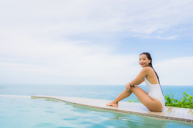 La giovane donna asiatica del ritratto si rilassa il sorriso felice intorno alla piscina all'aperto nella località di soggiorno dell'hotel con la vista di oceano del mare