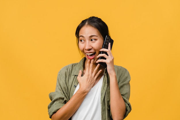 La giovane donna asiatica parla al telefono con espressione positiva, sorride ampiamente, vestita con abiti casual sentendosi felice e sta isolata sul muro giallo. La donna felice adorabile felice si rallegra del successo.