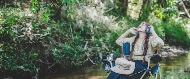 La giovane donna asiatica si siede su una sedia vicino al ruscello ascoltando musica con le cuffie wireless e usa il tablet felicemente mentre si accampa nello spazio della copia del bosco