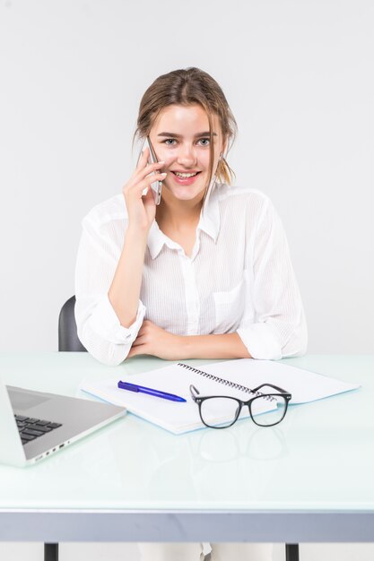 La giovane donna attraente in abbigliamento casual pastello che parla sul telefono cellulare, si siede il lavoro allo scrittorio con il computer portatile del pc isolato su fondo grigio.