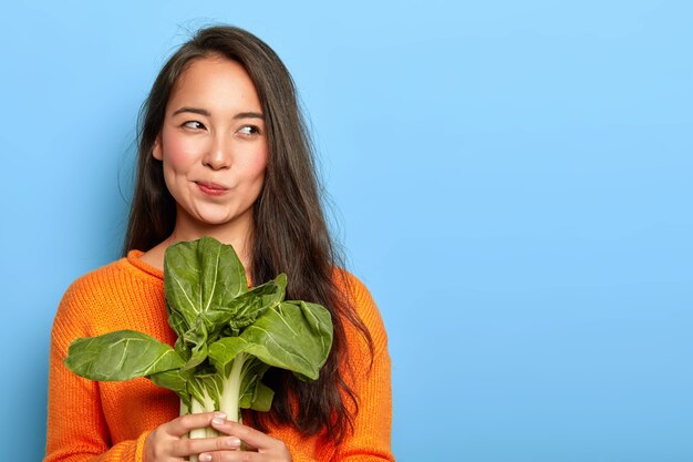 La giovane donna attraente tiene la verdura fresca verde, mangia cibo sano a casa, utilizza prodotti alimentari per preparare insalata vegetariana, indossa un maglione arancione, posa al coperto