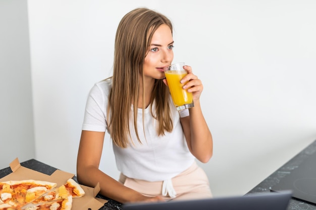 La giovane donna beve succo di frutta e mangia pizza e guarda il laptop in cucina