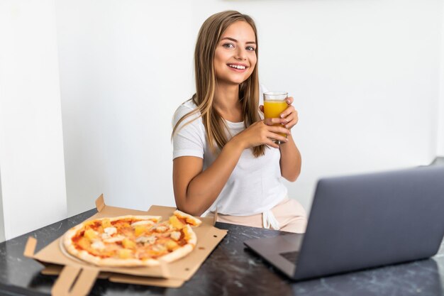 La giovane donna beve succo di frutta e mangia pizza e guarda il laptop in cucina