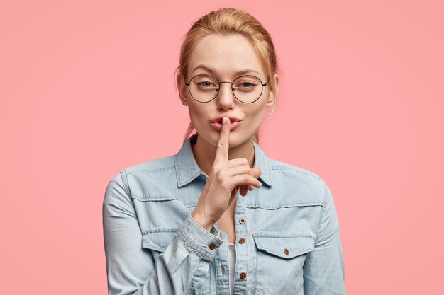 La giovane donna bionda adorabile sicura mostra il segno di silenzio, dice il segreto a qualcuno, tocca il dito anteriore sulle labbra