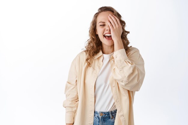 La giovane donna bionda felice copre la metà del viso ridendo e sorridendo spensierata in piedi in abiti casual su sfondo bianco