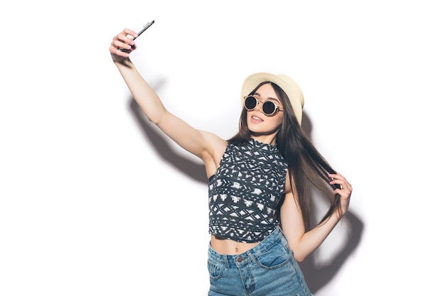 La giovane donna castana attraente allegra sta sorridendo sul muro bianco prendendo selfie con il suo telefono, indossando abiti estivi casual e un cappello