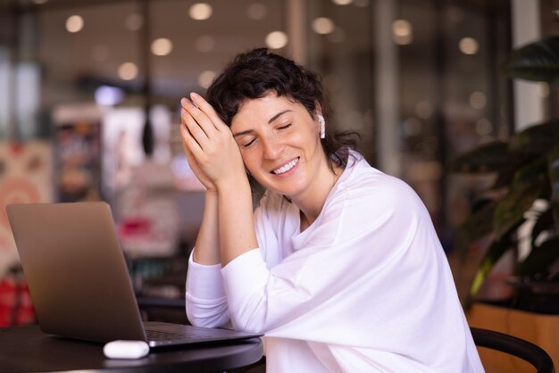 La giovane donna castana caucasica rilassata in maglione bianco che chiude gli occhi sta riposando durante la pausa dal lavoro al computer portatile nella caffetteria