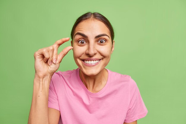 La giovane donna castana felice mostra le forme minuscole o piccole di gesto con le dita indossa la maglietta rosa casuale isolata sopra la parete verde