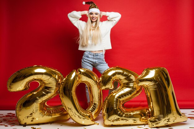 La giovane donna caucasica allegra in maglione bianco, jeans blu e cappello rosso di natale tiene le mani vicino alla testa, immagine isolata su sfondo rosso