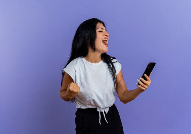 La giovane donna caucasica allegra tiene il telefono e tiene il pugno che guarda il lato