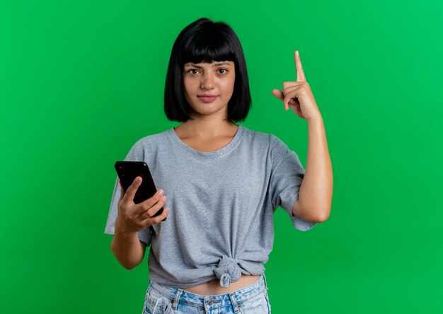 La giovane donna caucasica castana confusa tiene il telefono e indica in alto isolato su priorità bassa verde con lo spazio della copia