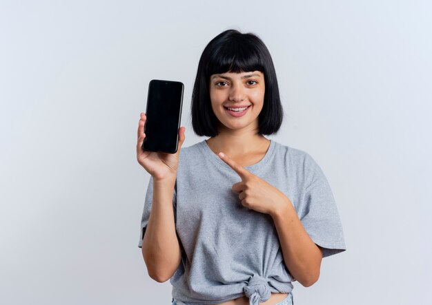 La giovane donna caucasica castana sorridente tiene e indica il telefono