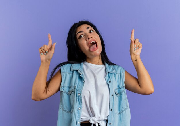 La giovane donna caucasica divertente attacca la lingua fuori e indica con due mani isolate su fondo viola con lo spazio della copia