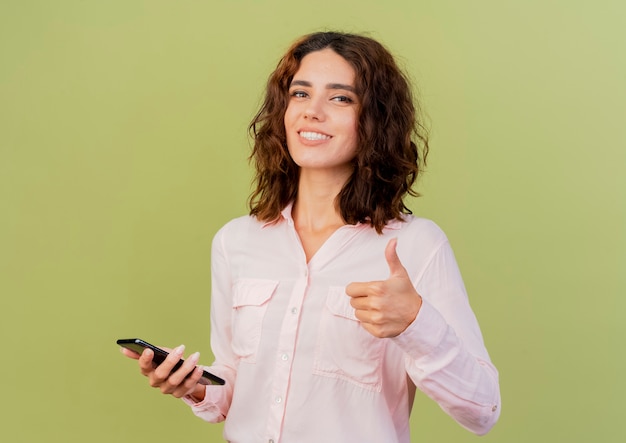 La giovane donna caucasica sorridente tiene il telefono e il pollice in alto isolato su priorità bassa verde con lo spazio della copia