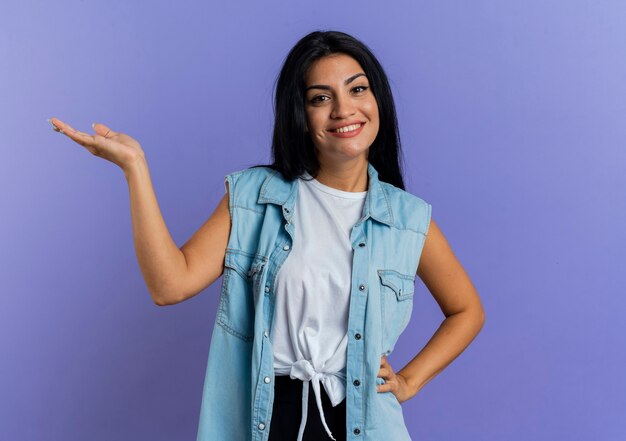 La giovane donna caucasica sorridente tiene la mano aperta isolata su fondo viola con lo spazio della copia