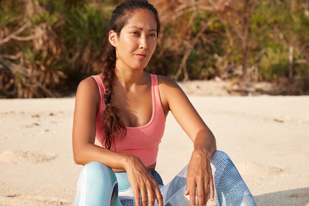 La giovane donna che fa jogging in abbigliamento sportivo si sente in salute, guarda pensieroso in lontananza, posa sulla spiaggia sabbiosa