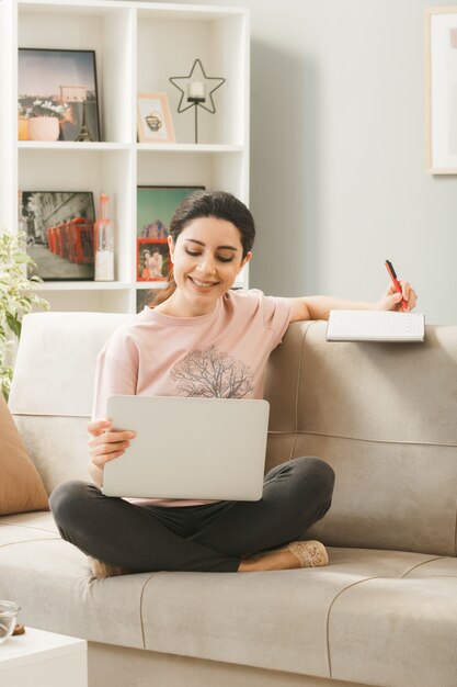 La giovane donna che si siede sul sofà dietro il tavolino da caffè che tiene e il computer portatile usato scrive sul libro in soggiorno