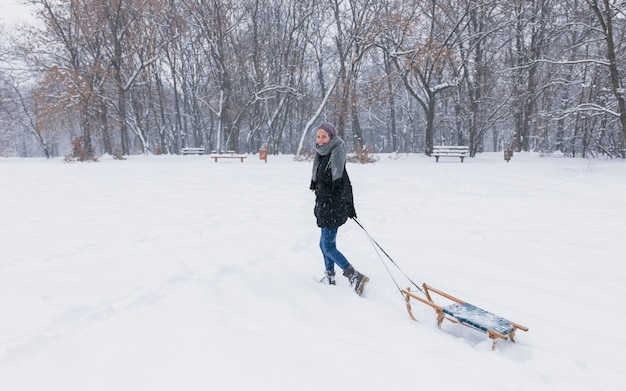 La giovane donna che tira la slitta di legno vuota su neve abbellisce alla foresta