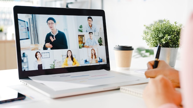 La giovane donna d'affari asiatica che utilizza il laptop parla con un collega del piano in una riunione di videochiamata mentre lavora da casa in soggiorno.