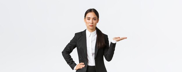 La giovane donna d'affari asiatica determinata dall'aspetto serio presenta il suo progetto mostrando qualcosa sul palmo che tiene la mano proprio su uno spazio vuoto mentre introduce il prodotto in piedi in vestito nero su sfondo bianco