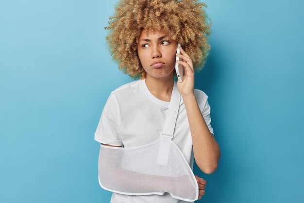 La giovane donna dai capelli ricci dispiaciuta fa una telefonata al medico ha il braccio rotto avvolto in una stecca vestita con una maglietta bianca casual distoglie lo sguardo pone su sfondo blu Problemi di salute