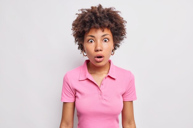 La giovane donna dai capelli ricci scioccata con i capelli afro fissa stordita sul davanti indossa una maglietta rosa casual tiene la bocca aperta sembra incredula isolata sul muro bianco