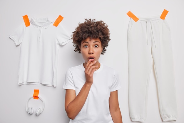 La giovane donna dai capelli ricci scioccata sembra stupita tiene la bocca aperta vestita con una t-shirt casual basic posa su sfondo bianco dello studio con pantaloni t-shirt intonacati e cuffie Omg concept