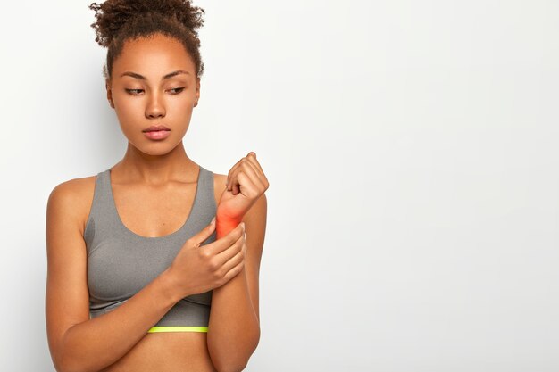 La giovane donna dai capelli ricci seria ha un polso infortunato, alza la mano con il fuoco rosso che mostra la posizione del dolore, soffre di distorsione dopo l'allenamento, indossa un top casual, posa sul muro bianco con lo spazio della copia