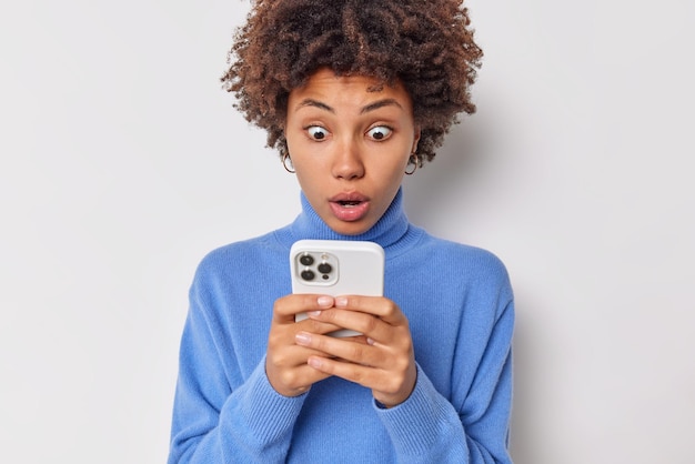 La giovane donna dai capelli ricci stupefatta fissa lo smartphone con un'espressione scioccata ha ricevuto un messaggio inaspettato dal fidanzato formale che reagisce a notizie sorprendenti indossa un dolcevita blu isolato su un muro bianco