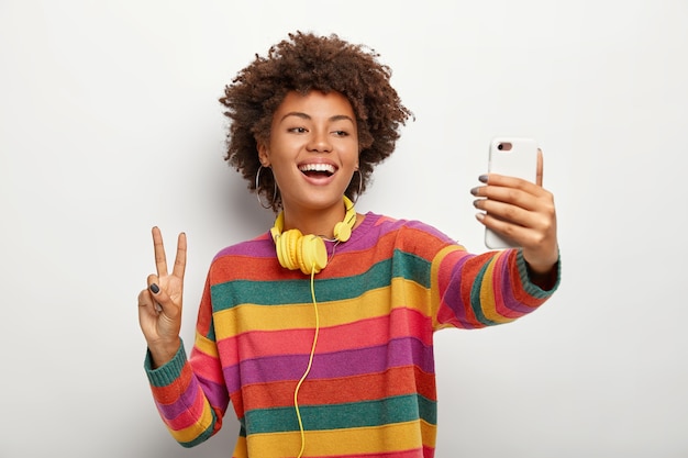 La giovane donna dai capelli riccia spensierata prende il ritratto del selfie sul cellulare, mostra il gesto di pace, indossa il maglione colorato a strisce