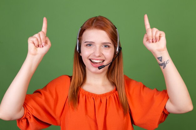 La giovane donna dai capelli rossi con una camicetta arancione casual sul call center del call center del manager della parete verde aiuta il lavoratore in linea a sorridere punta le dita