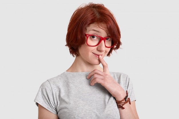 La giovane donna dai capelli rossi curiosa tiene il dito anteriore sulle labbra, guarda con interesse, indossa una maglietta casual con gli occhiali cerchiati rossi, sogna qualcosa, ha una pettinatura corta, isolata sul muro bianco.