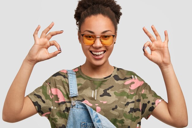 La giovane donna dalla pelle scura soddisfatta tiene le mani nel gesto giusto, vestita con una maglietta mimetica casual, una tuta di jeans