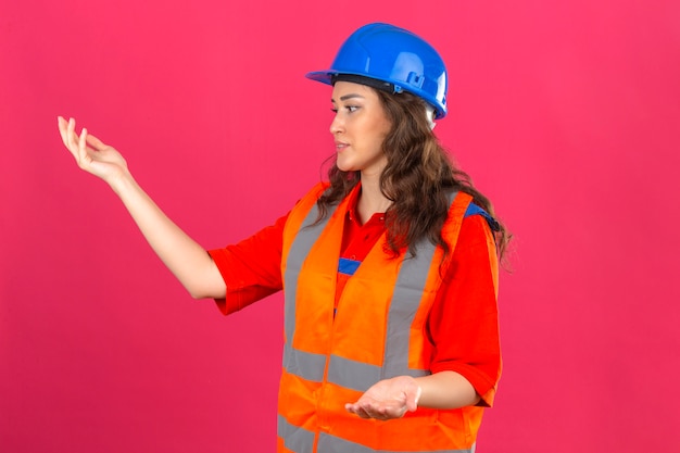 La giovane donna del costruttore in uniforme della costruzione e casco di sicurezza che gesturing la delusione stante con le mani si sono alzate sopra la parete rosa isolata