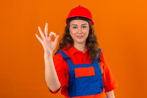 La giovane donna del costruttore in uniforme della costruzione e casco di sicurezza che sembrano sorridere sicuro allegramente che fa bene firmano firmano sopra la parete arancio isolata