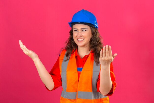 La giovane donna del costruttore in uniforme della costruzione e casco di sicurezza che solleva le mani lateralmente con sgomento e lo sguardo di delusione confuso ha confuso che cosa è accaduto sopra la parete rosa isolata