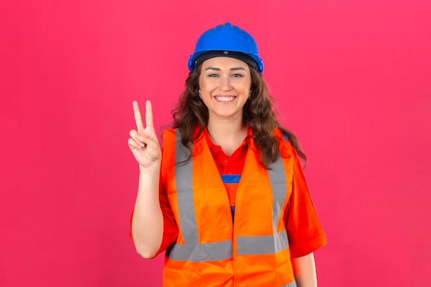 La giovane donna del costruttore in uniforme della costruzione e casco di sicurezza che sorridono allegramente e che fanno la vittoria firmano sopra la parete rosa isolata