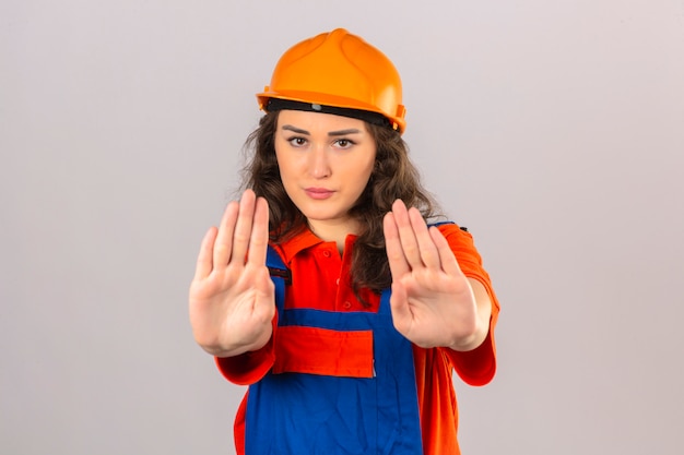 La giovane donna del costruttore nell'uniforme della costruzione e nel casco di sicurezza che fanno l'arresto cantano con le palme dell'espressione d'avvertimento delle mani sopra la parete bianca isolata