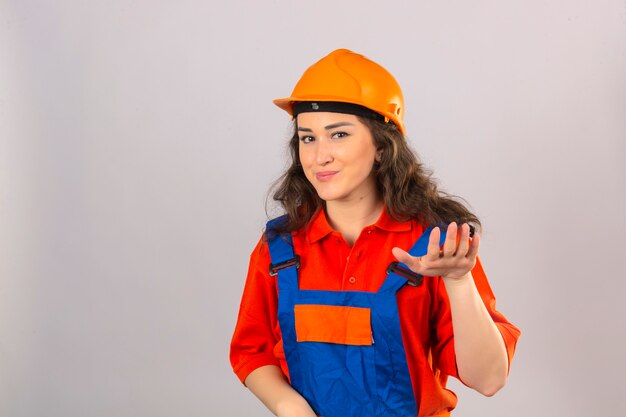 La giovane donna del costruttore nell'uniforme della costruzione e nel chiedere il casco di sicurezza si rilassa prendono il gesto facile di calma della tenuta della mano che sorride amichevole sopra la parete bianca isolata