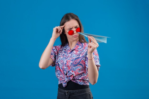 La giovane donna del viaggiatore che indossa gli occhiali da sole rossi che tengono l'aeroplano di carta che lo esamina con il fronte aggrottante le sopracciglia ha dispiaciuto avere dubbi sulla parete blu