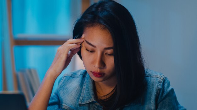 La giovane donna di affari cinese millenaria che lavora a tarda notte si affanna con il problema di ricerca di progetto sul computer portatile in salone a casa moderna. Concetto di sindrome di burnout professionale delle persone dell'Asia.