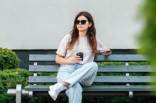 La giovane donna di affari si siede su una panchina con una tazza di caffè e uno smartphone