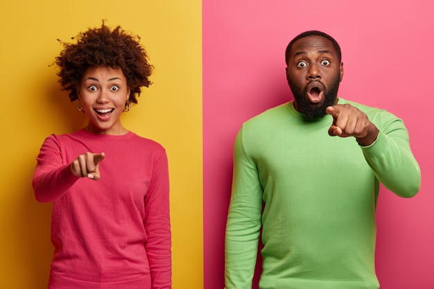 La giovane donna e l'uomo afroamericani eccitati indicano davanti, indicano con espressioni sorprese, indossano abiti luminosi, si sentono imbarazzati, posano su due pareti colorate. Wow, guarda là