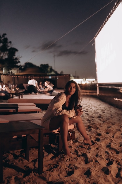 La giovane donna è seduta sulla sedia in spiaggia di notte e guardando la fotocamera