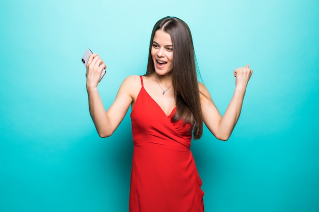 La giovane donna eccitata in vestito rosso fa il gesto del vincitore in piedi isolato sopra la parete blu