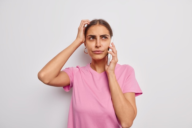 La giovane donna esitante e perplessa si gratta la testa cerca di ricordare qualcosa in mente ha una conversazione telefonica vestita casualmente isolata su un muro bianco