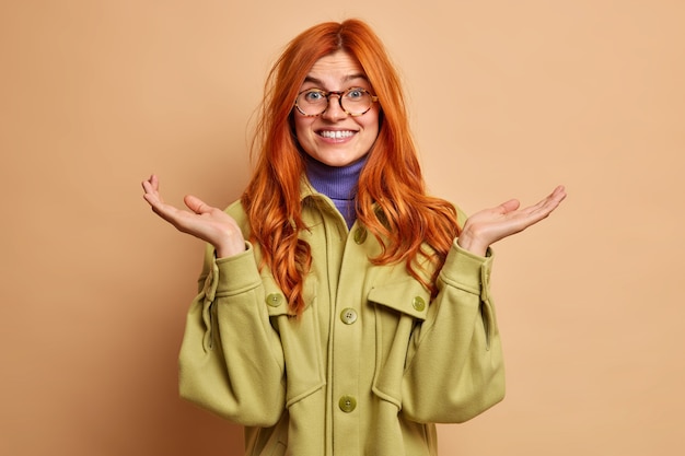 La giovane donna europea allegra confusa ignara con i capelli rossi alza i palmi ha un'espressione dubbiosa non può rispondere immediatamente indossa la giacca verde.